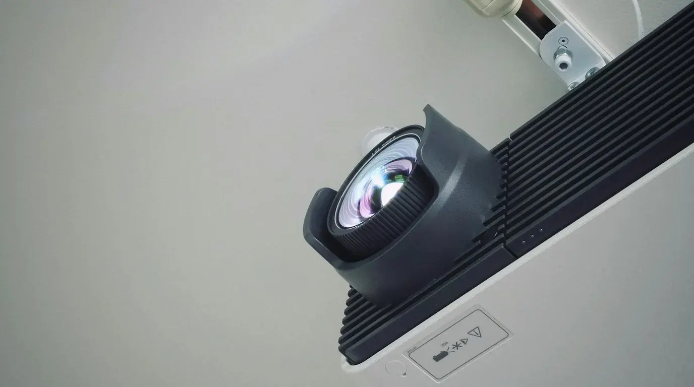 A ceiling-mounted projector seen from below, its lens catching the light against a white ceiling