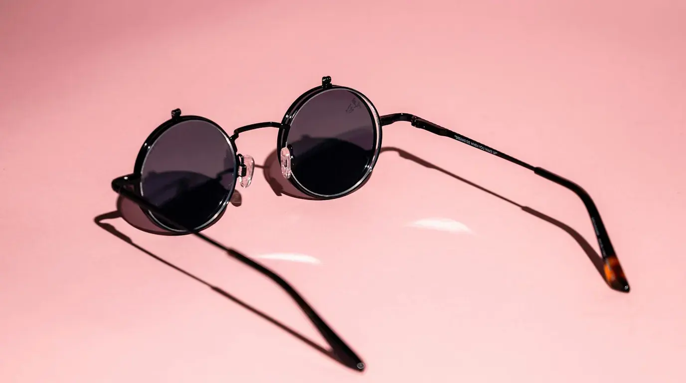 A pair of round black sunglasses lying open on a plain pink studio backdrop, casting long shadows